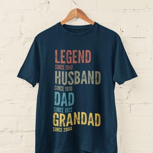 May include: A navy blue short-sleeved t-shirt featuring distressed, multi-coloured text that reads: LEGEND SINCE 1947, HUSBAND SINCE 1970, DAD SINCE 1972, and GRANDAD SINCE 2000. The shirt is displayed on a wooden hanger against a white brick wall.