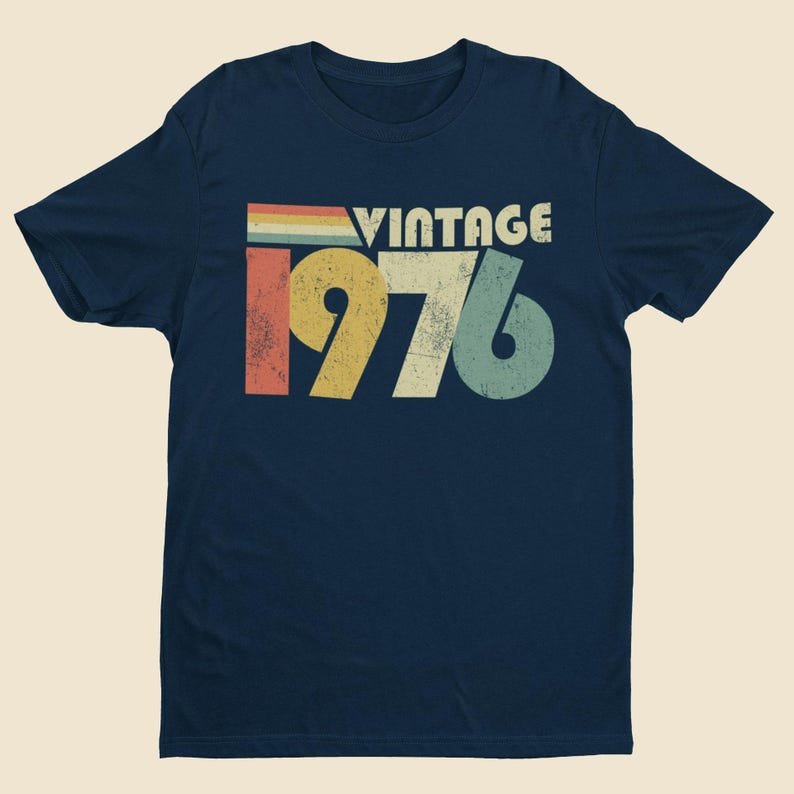 May include: A navy blue short-sleeved t-shirt featuring a distressed graphic print on the chest. The design includes the word VINTAGE in white letters above the large numbers 1976, rendered in yellow, white, and teal with a retro striped accent.