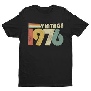 May include: A black short-sleeve t-shirt featuring a graphic print with the text VINTAGE 1976. The design includes a distressed, multi-colored horizontal stripe pattern above the year, rendered in muted shades of red, yellow, and teal blue.