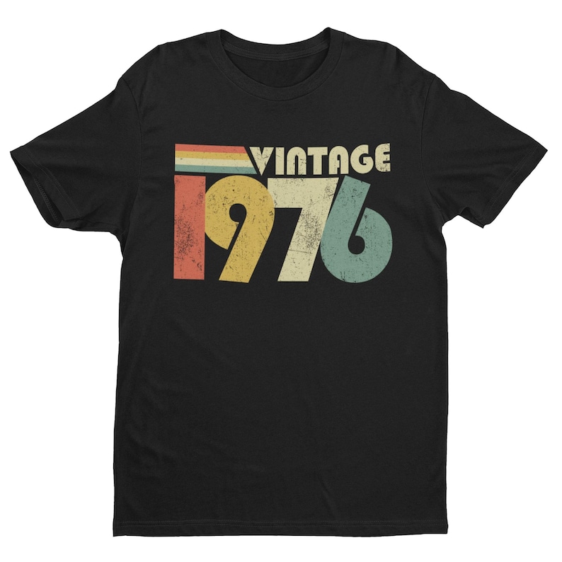 May include: A black short-sleeve t-shirt featuring a graphic print with the text VINTAGE 1976. The design includes a distressed, multi-colored horizontal stripe pattern above the year, rendered in muted shades of red, yellow, and teal blue.