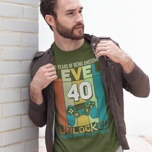 Op de afbeelding: Een groen T-shirt met een retro-grafisch ontwerp ter ere van een 40e verjaardag. Het ontwerp bevat een videogamecontroller, het nummer 40 en de tekst "Level 40 Unlocked Press Start to Continue".