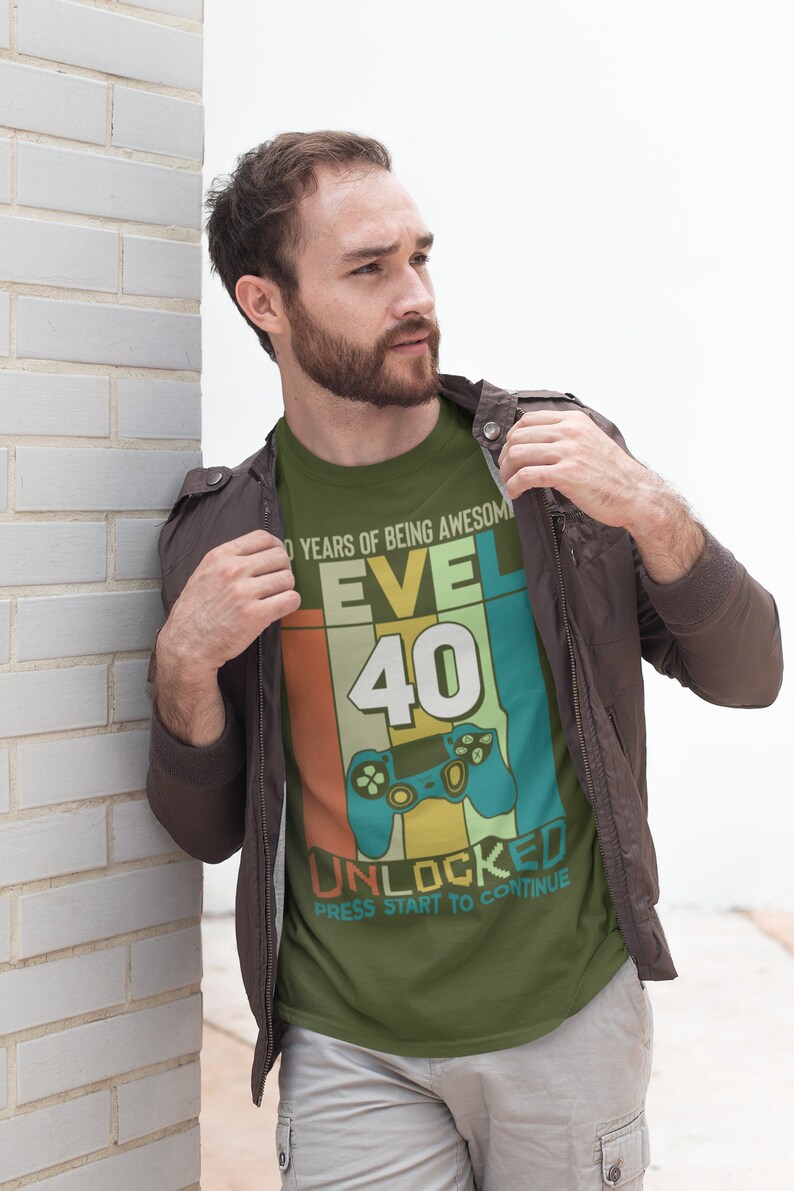 Op de afbeelding: Een groen T-shirt met een retro-grafisch ontwerp ter ere van een 40e verjaardag. Het ontwerp bevat een videogamecontroller, het nummer 40 en de tekst "Level 40 Unlocked Press Start to Continue".