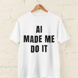 Op de afbeelding: Wit T-shirt met de tekst "AI MADE ME DO IT" in vette zwarte letters. Het shirt hangt aan een houten hanger tegen een witte bakstenen muur. Het T-shirt is een casual kledingstuk.