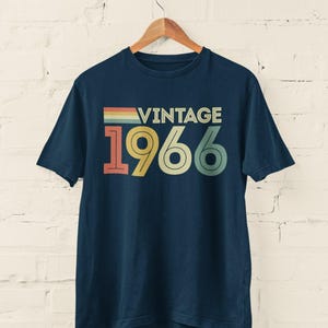 Può includere: Una t-shirt a maniche corte blu navy con la parola VINTAGE in un carattere sans-serif sopra i grandi numeri 1966. Il design include una barra orizzontale a strisce multicolori e utilizza una tavolozza di colori retrò in rosso, giallo e verde.