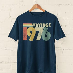 May include: A navy blue short-sleeve t-shirt featuring a graphic print on the chest. The design includes the word VINTAGE in white letters above the large numbers 1976, which are rendered in distressed red, yellow, and teal colors.