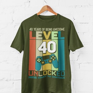 Op de afbeelding: Groen T-shirt met een retro-stijl grafische afbeelding met de tekst "40 Years of Being Awesome Level 40 Unlocked Press Start to Continue". De afbeelding bevat een videogamecontroller en een regenboogkleurenschema.