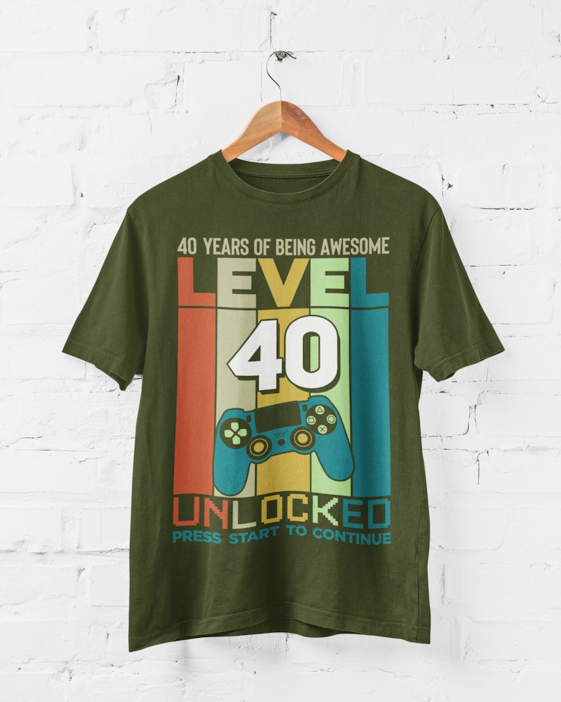 Op de afbeelding: Groen T-shirt met een retro-stijl grafische afbeelding met de tekst "40 Years of Being Awesome Level 40 Unlocked Press Start to Continue". De afbeelding bevat een videogamecontroller en een regenboogkleurenschema.