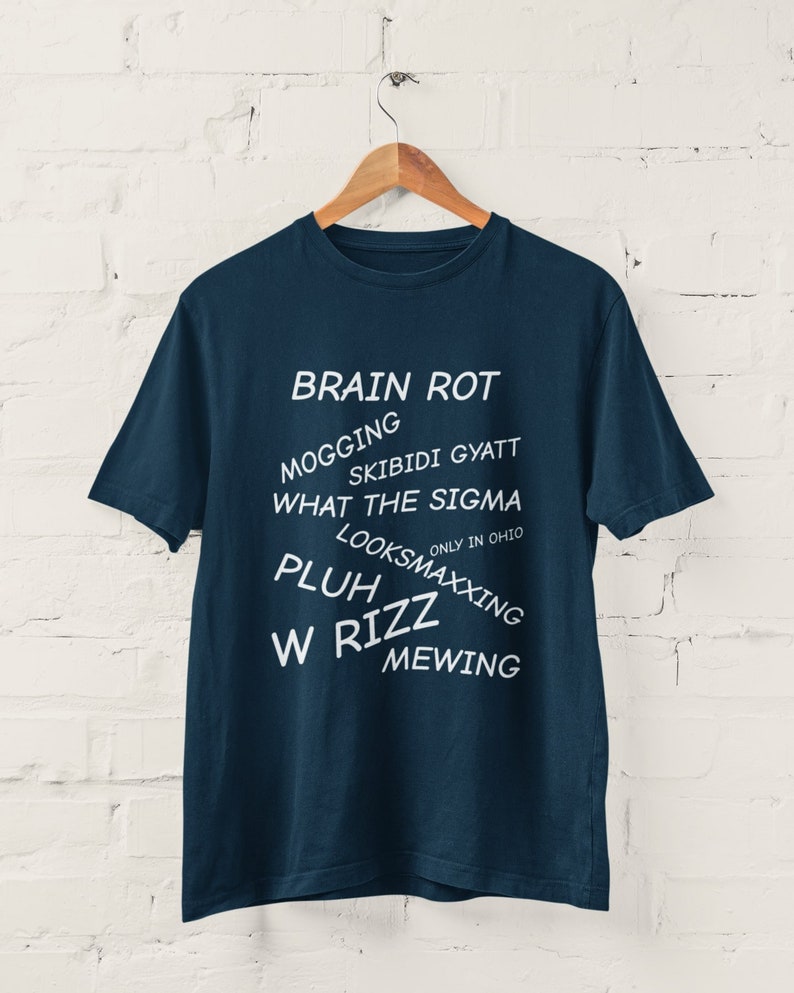 May include: A navy blue t-shirt with white text that reads "BRAIN ROT", "MOGGING", "SKIBIDI GYATT", "WHAT THE SIGMA", "ONLY IN OHIO", "LOOKSMAXXING", "PLUH", "W RIZZ", and "MEWING".
