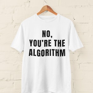 Może przedstawiać: Biała koszulka z napisem "NO, YOU'RE THE ALGORITHM" pogrubionymi, czarnymi literami. Koszulka wisi na drewnianym wieszaku na tle białej ceglanej ściany.
