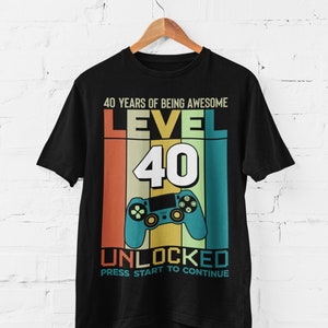 Op de afbeelding: Zwart T-shirt met een retro-grafisch ontwerp ter ere van een 40e verjaardag. Het ontwerp bevat een videogamecontroller, de tekst "40 Years of Being Awesome", "Level 40" en "Unlocked". De tekst "Press Start to Continue" is ook inbegrepen.