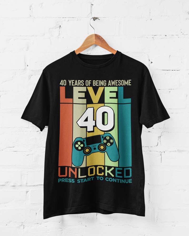 Op de afbeelding: Zwart T-shirt met een retro-grafisch ontwerp ter ere van een 40e verjaardag. Het ontwerp bevat een videogamecontroller, de tekst "40 Years of Being Awesome", "Level 40" en "Unlocked". De tekst "Press Start to Continue" is ook inbegrepen.