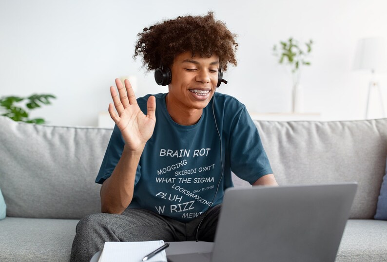 May include: A young person wearing a blue t-shirt with the text "BRAIN ROT MOGGING SKIBIDI GYATT WHAT THE SIGMA LOOKSMAXING PLUH W RIZZ MEW" is sitting on a couch and waving at a laptop screen.