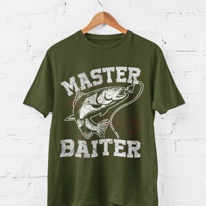 May include: An olive green short-sleeve t-shirt featuring a white distressed graphic of a fish on a fishing line. The text "MASTER BAITER" is printed in a bold, weathered white font above and below the fish illustration on the front.