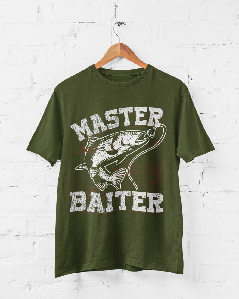 May include: An olive green short-sleeve t-shirt featuring a white distressed graphic of a fish on a fishing line. The text "MASTER BAITER" is printed in a bold, weathered white font above and below the fish illustration on the front.