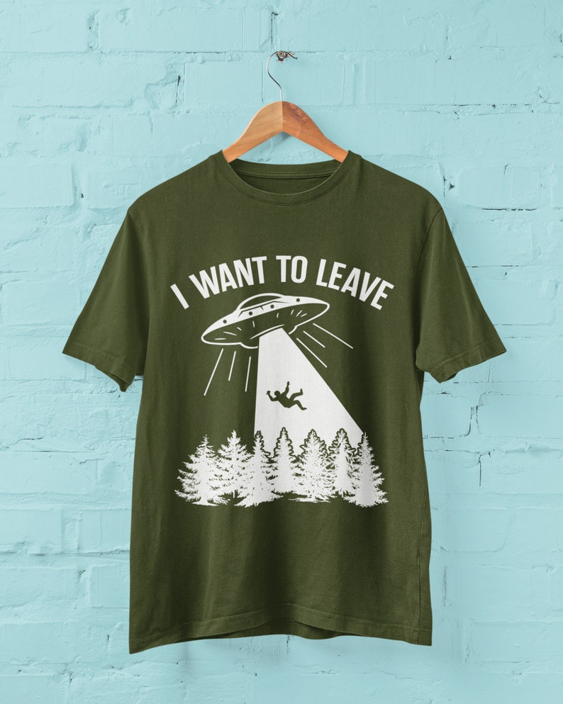 May include: A green t-shirt with a white graphic of a UFO and a person falling from the sky. The text "I WANT TO LEAVE" is printed on the shirt.