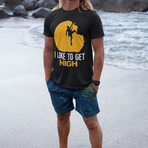 May include: A man wearing a black t-shirt with a yellow circle and a silhouette of a rock climber. The text "I LIKE TO GET HIGH" is printed on the shirt. He is standing on a sandy beach with the ocean in the background.