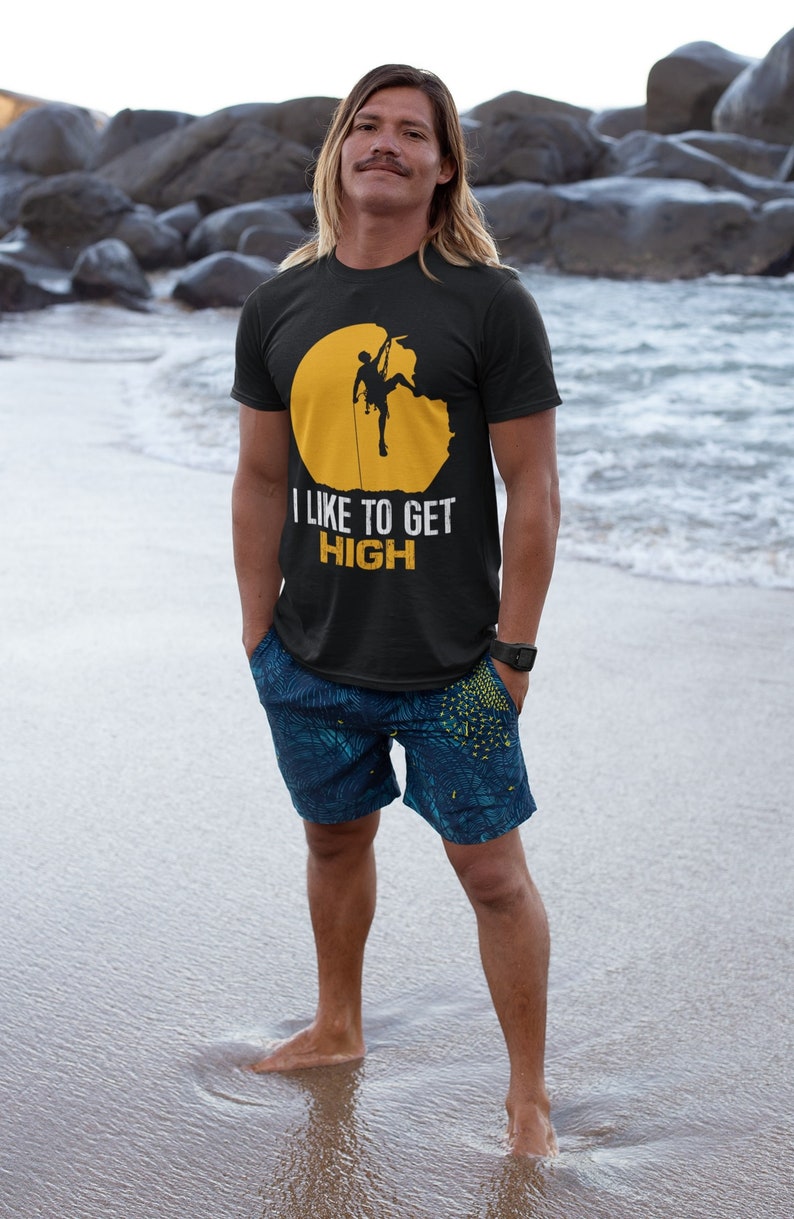 May include: A man wearing a black t-shirt with a yellow circle and a silhouette of a rock climber. The text "I LIKE TO GET HIGH" is printed on the shirt. He is standing on a sandy beach with the ocean in the background.