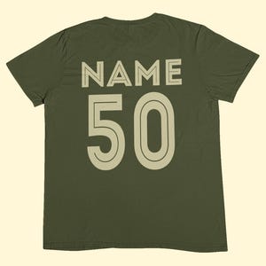 May include: An olive green short-sleeve t-shirt featuring the word NAME printed in a light beige sans-serif font above the number 50 in a matching light beige font. The design is centered on the back of the garment with a clean, bold aesthetic.