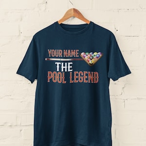 May include: A navy blue t-shirt with the text "YOUR NAME THE POOL LEGEND" in orange and white. A pool cue and a rack of billiard balls are pictured above the text.