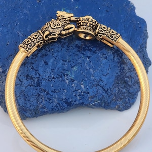 May include: Gold-colored bangle bracelet with ornate detailing. The bracelet features a textured band and decorative clasps with intricate patterns and a small blue accent. The clasp design includes animal-like figures.