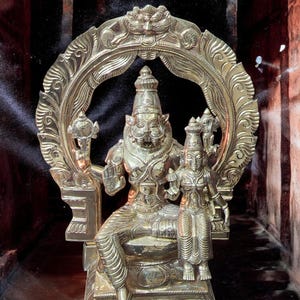 May include: A detailed brass sculpture of a deity seated on a throne, with an ornate halo. The figure has a lion-like face and is accompanied by a smaller figure. The sculpture is set against a dark background.