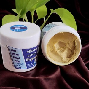May include: Two white plastic jars of Gopi Tilak, a quality product from India. One jar is closed, displaying a blue and white label with text in English and Hindi. The other jar is open, revealing a light brown paste. Green leaves are in the background.