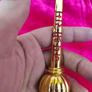 Op de afbeelding: Een goudkleurig metalen voorwerp met een ronde, bolvormige basis en een lange, cilindrische bovenkant. De bovenkant heeft een kleine, heldere edelsteen aan het einde en rode letters die "श्री हनुमान" lezen.