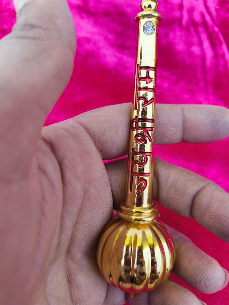 Op de afbeelding: Een goudkleurig metalen voorwerp met een ronde, bolvormige basis en een lange, cilindrische bovenkant. De bovenkant heeft een kleine, heldere edelsteen aan het einde en rode letters die "श्री हनुमान" lezen.