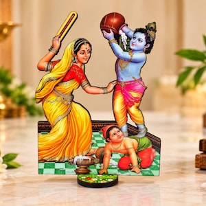 May include: A decorative cutout display featuring a woman in a yellow sari holding a wooden tool, a blue-skinned figure pouring liquid from a brown pot, and a small child crawling on a green and white checkered floor with spilled white liquid.