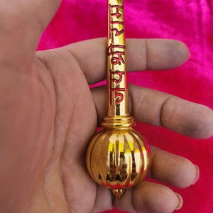Op de afbeelding: Een gouden metalen ceremoni&euml;le knots met een ronde, bolvormige kop en een lange, cilindrische steel. De steel is beschreven met rode letters in een onbekende taal.