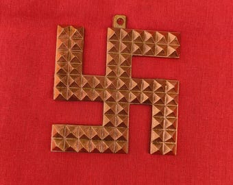 Puede incluir: Un colgante de esvástica de color cobre con una superficie texturizada. El colgante está hecho de metal y tiene un pequeño bucle en la parte superior para colgar.