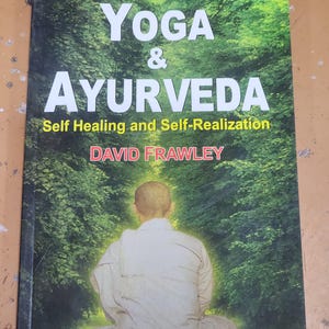 May include: A book titled "Yoga & Ayurveda: Self Healing and Self-Realization" by David Frawley. The cover features a person sitting in a meditative pose in a lush green forest.