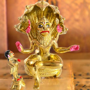 May include: A gold-colored statue of a deity with a crown-like headdress and multiple arms, seated with a smaller figure in front. The deity has pink accents on the hands and ears. The statue is on a gold-colored surface.