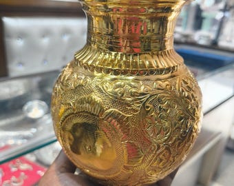 May include: A gold-colored, ornate metal vase with intricate embossed floral patterns. The vase has a rounded body and a flared rim. The surface is highly reflective, and the vase is held in a person's hand.