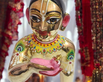 Könnte beinhalten: Goldene Statue einer Gottheit mit schwarzem Haar und goldenem Gesicht. Die Statue hält eine rosa Blume in den Händen und ist mit farbenfrohen Details verziert, darunter Pfauenfedermuster auf den Armen und ein dekorativer Kragen.