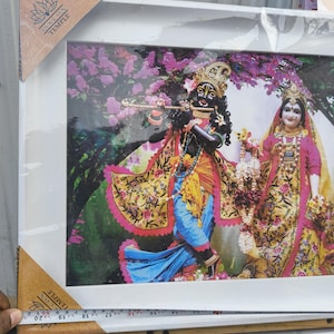 May include: A framed print of a Hindu deity couple, Krishna and Radha, in colorful clothing with floral patterns. The frame is white with a brown wooden border. The print is surrounded by a pink and white floral background.