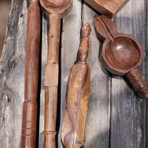 Puede incluir: Colección de utensilios de cocina de madera hechos a mano, que incluyen un rodillo, cucharas y una prensa cuadrada. Los artículos están hechos de madera marrón oscura y están dispuestos sobre una superficie de madera envejecida.