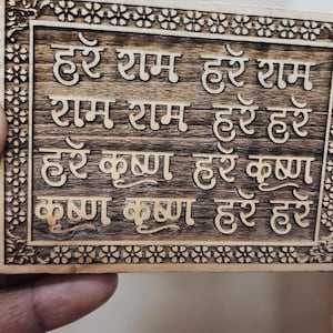 May include: Wooden plaque with carved Sanskrit text. The text is in a light color, set against a dark brown background. The plaque has a decorative floral border.