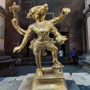 May include: A gold statue of a deity with multiple arms and a pig-like head. The statue stands on a square base and holds a circular object. The background shows an ancient building.
