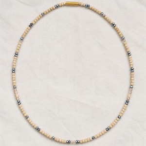 Puede incluir: Un collar de cuentas con un patrón repetitivo de cuentas cilíndricas de color tostado claro intercaladas con pequeñas cuentas metálicas de color gris oscuro facetadas y cuentas transparentes, con un cierre magnético cilíndrico dorado.