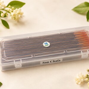 May include: A rectangular, clear plastic storage box containing dark brown incense sticks. The lid features a small circular logo with the letters SPP and a green leaf graphic. A white label on the front of the box displays the text Son Chafa.