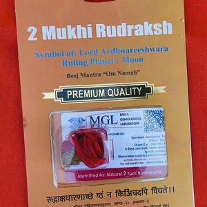 May include: A red string tied around a brown and red Rudraksha bead. The bead is labeled as a "Natural 2 Face Rudraksha" and is certified by the Maya Gemological Laboratory. The card also includes the text "2 Mukhi Rudraksha", "Symbol of: Lord Ardhnareeshwara", "Ruling Planet: Moon", "Beej Mantra "Om Namah"", and "PREMIUM QUALITY".