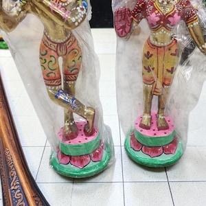Puede incluir: Dos estatuas doradas de figuras con vestimenta tradicional, cada una sobre una base de flor de loto rosa y verde. Las figuras están adornadas con patrones coloridos y están encerradas en plástico. Un marco decorativo está en primer plano.