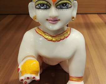 May include: A white figurine of a crawling deity with detailed facial features, including painted eyes, a yellow forehead marking, and a black hair bun. The figure has gold accents and is on a wooden surface.