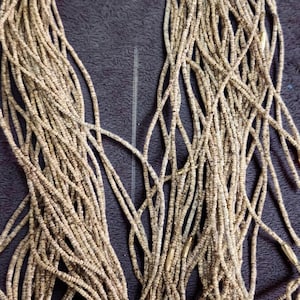 May include: A collection of beige and brown beaded necklaces. The beads are small and cylindrical, strung together to form long strands. The necklaces are arranged in a loose, intertwined pattern against a dark background.