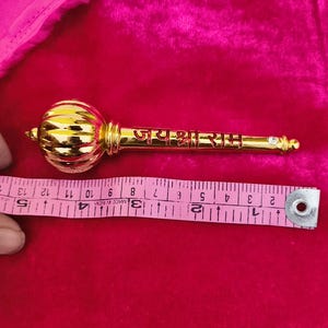 Op de afbeelding: Een gouden ceremoni&euml;le knots met een ronde knop aan de bovenkant en de woorden "जय श्री राम" gegraveerd op de schacht. De knots is ongeveer 30 cm lang.