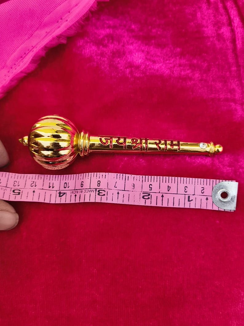 Op de afbeelding: Een gouden ceremoni&euml;le knots met een ronde knop aan de bovenkant en de woorden "जय श्री राम" gegraveerd op de schacht. De knots is ongeveer 30 cm lang.