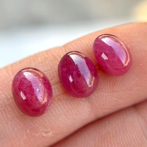 May include: Three oval-shaped, polished ruby gemstones. The gems are a deep, vibrant red with a glossy surface, reflecting light. The stones are displayed on a neutral background, highlighting their color and shape. These gemstones are suitable for jewelry making.