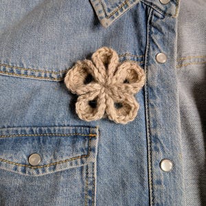 May include: A light beige, five-petaled crochet flower applique attached to a light blue denim shirt. The shirt features a pointed collar, a chest pocket, and silver-toned snap buttons. The crochet flower has a textured, yarn-based construction.