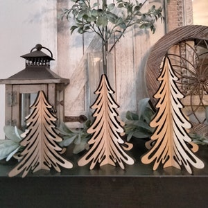 May include: Three wooden cutout Christmas trees in a light brown color. The trees are standing on a black surface in front of a white wooden background.